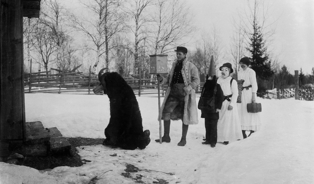 Fem utklädda personer som står på rad på snöig backe utanför stuga. 