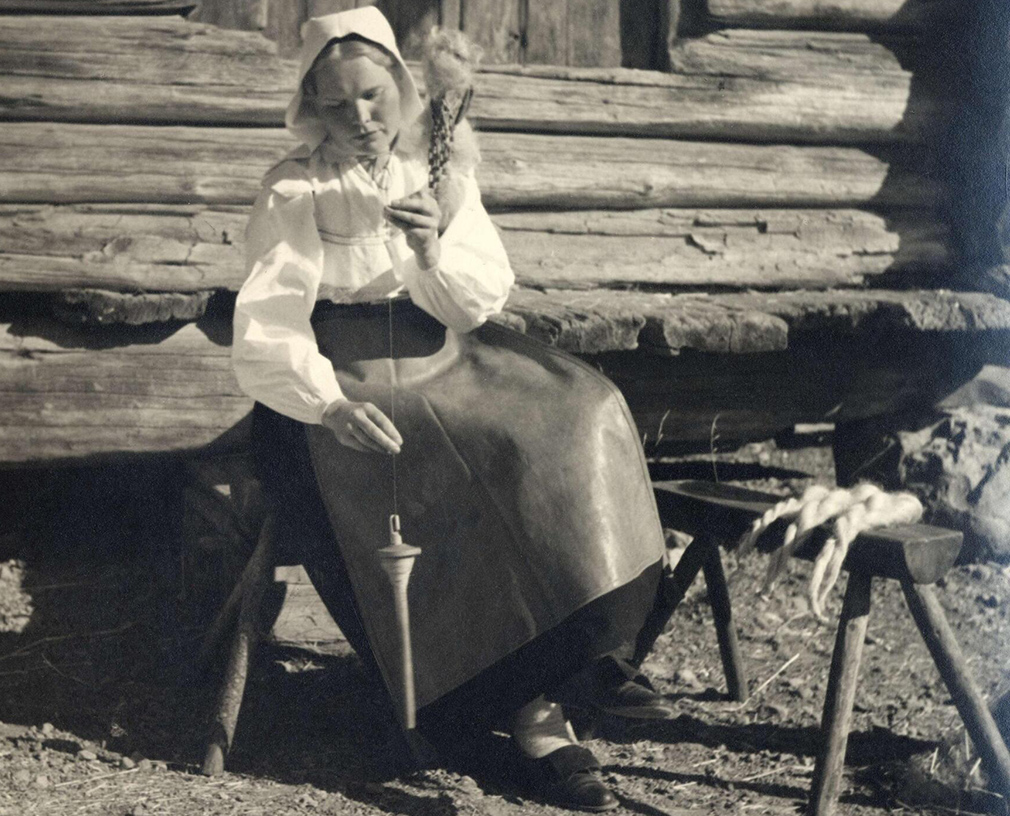 Ett svartvitt fotografi där en kvinna i folkdräkt sitter på en bänk utanför ett timmerhus och spinner garn med en "slända".